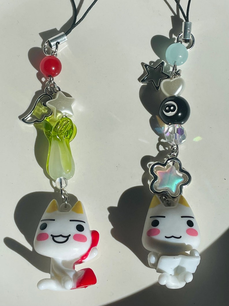 May include: Two white cat charm keychains with different accessories. One keychain has a green, white, and red charm with a star and a wing. The other keychain has a blue, white, and black charm with a star, a heart, and a black eight ball.