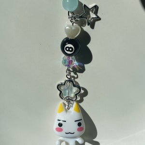 May include: White cat charm with yellow ears and pink cheeks, holding a blue and white fish. The charm is attached to a silver chain with a white heart, a black 8-ball, a blue crystal, a silver star, and a light blue bead.