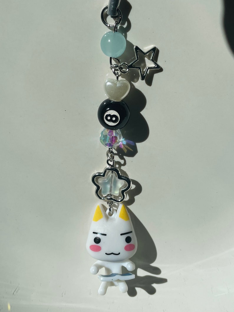 May include: White cat charm with yellow ears and pink cheeks, holding a blue and white fish. The charm is attached to a silver chain with a white heart, a black 8-ball, a blue crystal, a silver star, and a light blue bead.