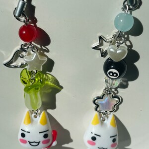 May include: Two white cat charm keychains with different accessories. The keychain on the left has a red bead, a green and white star, a green angel wing, and a green leaf. The keychain on the right has a blue bead, a silver star, a white heart, a black and white 8-ball, and a silver star with iridescent glitter.