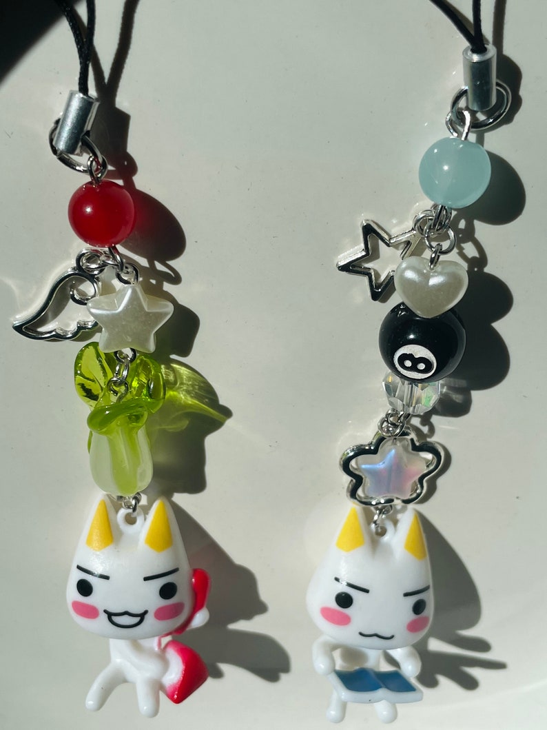 May include: Two white cat charm keychains with different accessories. The keychain on the left has a red bead, a green and white star, a green angel wing, and a green leaf. The keychain on the right has a blue bead, a silver star, a white heart, a black and white 8-ball, and a silver star with iridescent glitter.