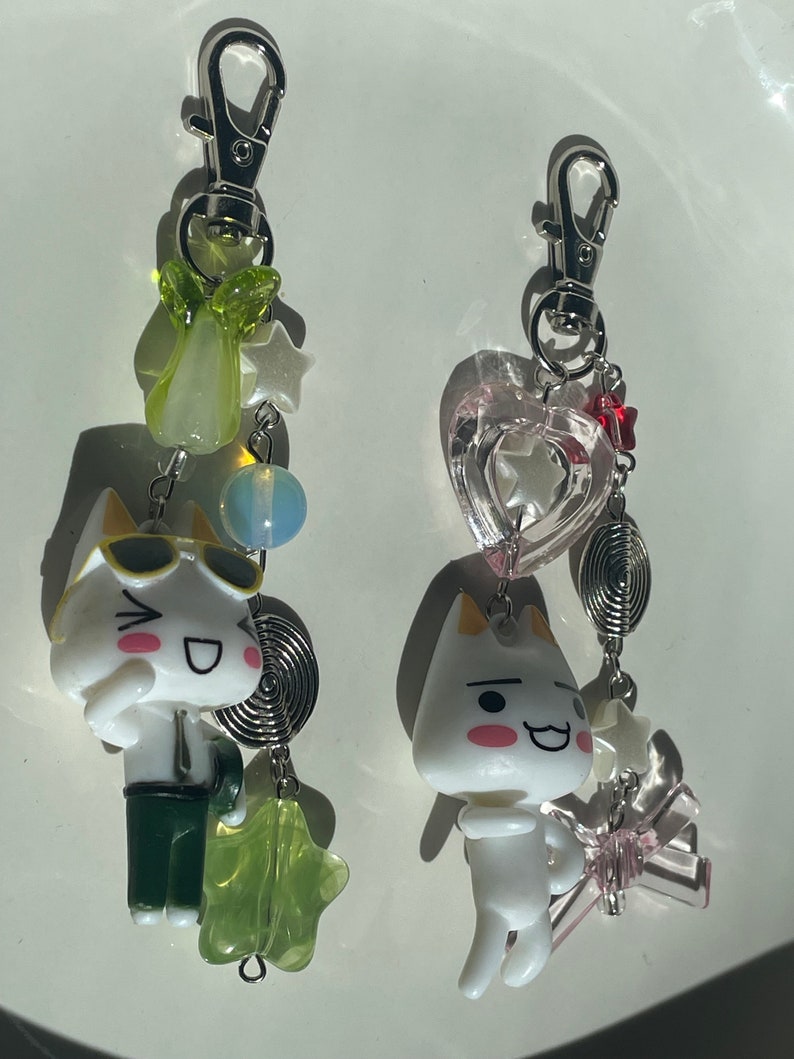 May include: Two white cat charm keychains with different accessories. One keychain has a green star, a green pear, a blue bead, and a silver spiral. The other keychain has a pink heart, a red star, a silver spiral, and a pink star.