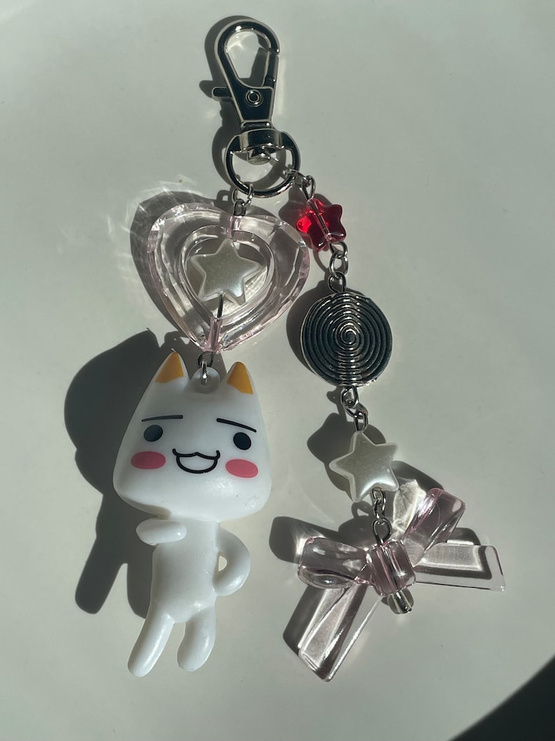 May include: A white cartoon cat keychain with a pink heart, a silver star, a silver spiral, a white star, and a pink bow charm.