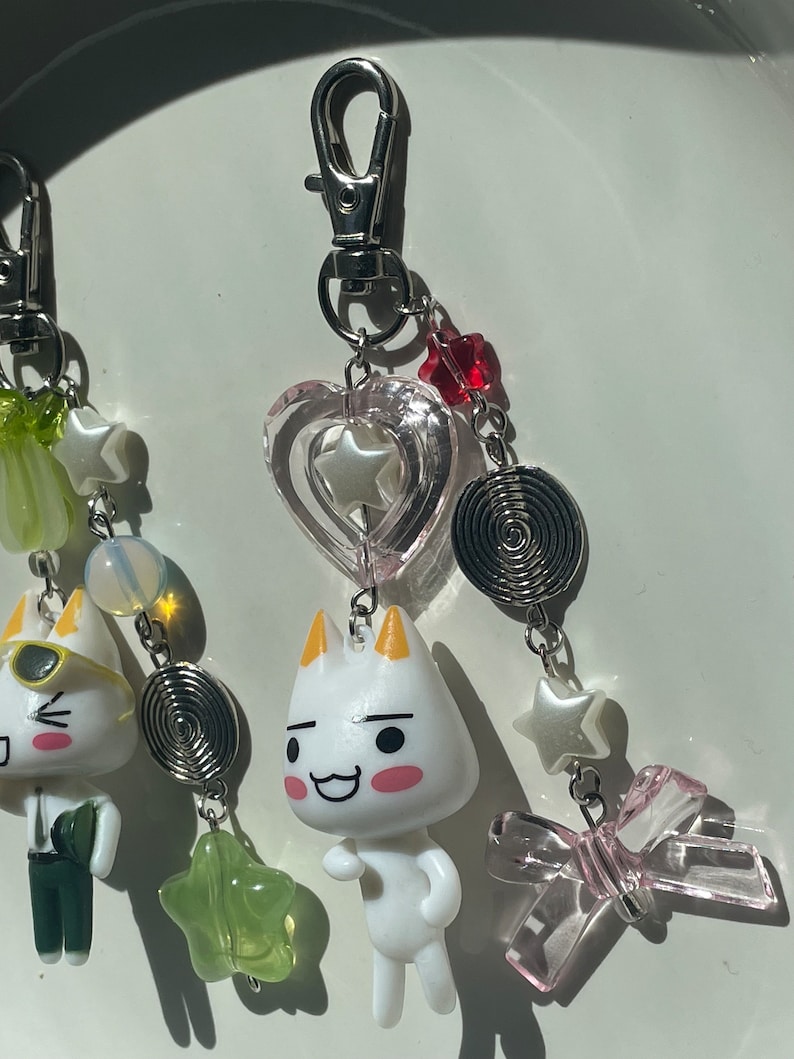 May include: A white cat charm with a pink heart, a green star, a silver spiral, and a pink bow. The charm is attached to a silver keychain.