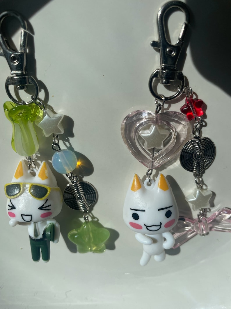 May include: Two white cartoon cat keychains with different expressions. One keychain has a green, yellow, and white charm with a star and a spiral. The other keychain has a pink heart charm with a star and a spiral.