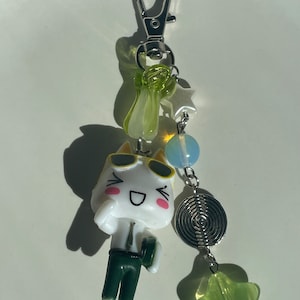 May include: A white cartoon cat keychain with green trousers and sunglasses. The keychain has a silver chain with green and blue beads, a silver star charm, and a silver spiral charm.