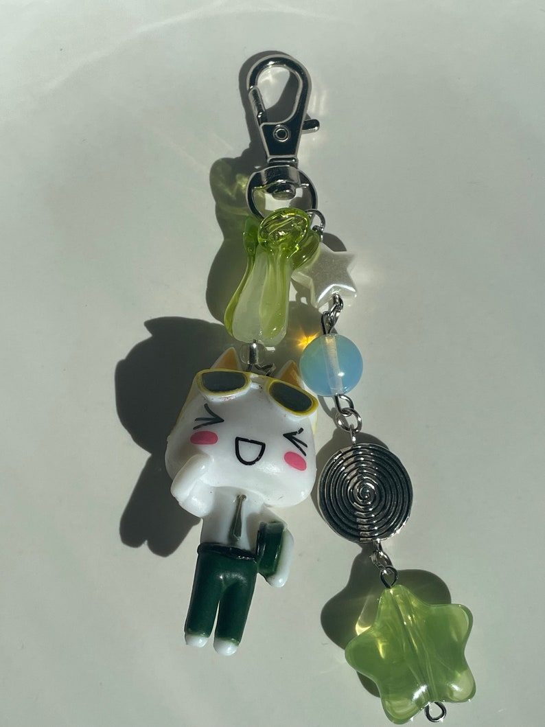 May include: A white cartoon cat keychain with green trousers and sunglasses. The keychain has a silver chain with green and blue beads, a silver star charm, and a silver spiral charm.