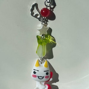 May include: A white cartoon cat charm with red and yellow accents, holding a red heart. The charm is attached to a silver chain with a green and white star, a silver wing, and a red bead.