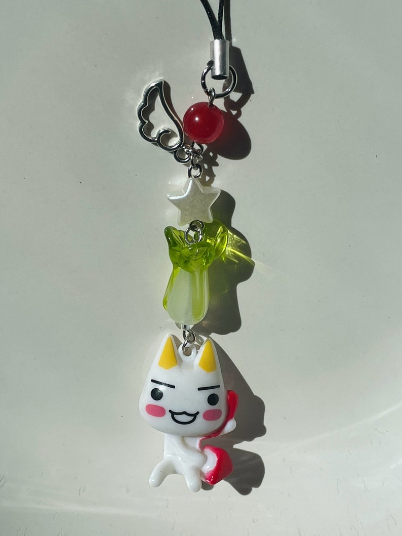 May include: A white cartoon cat charm with red and yellow accents, holding a red heart. The charm is attached to a silver chain with a green and white star, a silver wing, and a red bead.