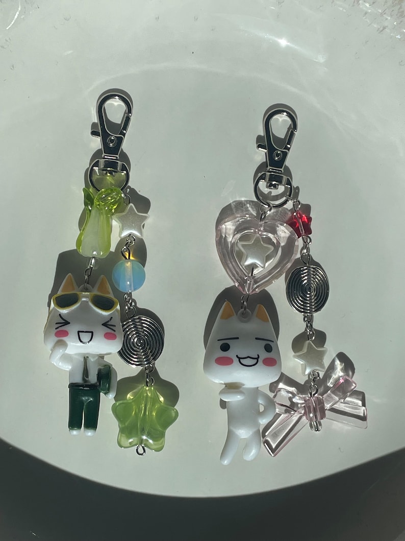 May include: Two white cat charm keyrings with different expressions. One cat is wearing sunglasses and a green outfit, the other is wearing a pink heart charm. Both charms have a silver spring and star charms.