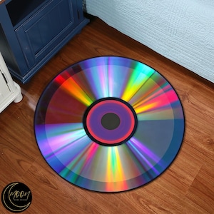 Op de afbeelding: Een rond tapijt dat is ontworpen om op een cd te lijken. Het tapijt heeft een levendig, iriserend kleurenschema met een centrale paarse cirkel en een zwarte rand. Het tapijt ligt op een houten vloer.