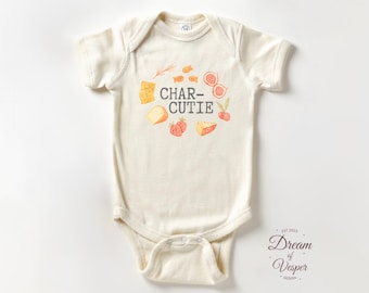 May include: Cream-colored baby onesie with the text "CHAR-CUTIE" in the center, surrounded by watercolor illustrations of cheese, figs, strawberries, and cherries. The onesie has short sleeves and a snap closure. The design includes the text "Dream of Vesper Design" and the year "est 2023".
