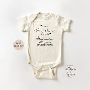 May include: Cream-colored baby onesie with black script text. The text reads "aunt Angelina & uncle Harvey will you be my godparents?" with heart accents. A small logo is in the bottom right corner.