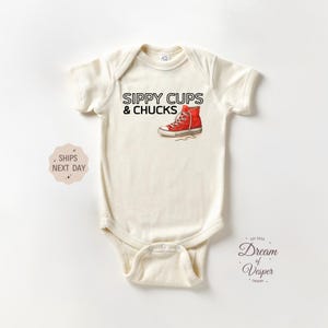 May include: Cream-colored baby bodysuit with the text "SIPPY CUPS & CHUCKS" and a red sneaker graphic. The bodysuit has short sleeves and a snap closure. The design includes the words "Dream of Vesper Design" and "Ships Next Day".