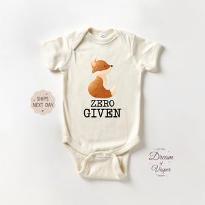 May include: Cream-colored baby bodysuit with a watercolor illustration of a fox and the words "ZERO GIVEN" in black. The fox is brown and white. The bodysuit is short-sleeved and has a snap closure.