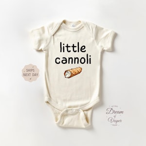 May include: Cream-colored baby onesie with the words "little cannoli" in black script. A cartoon illustration of a cannoli is below the text. The onesie has short sleeves and a snap closure.