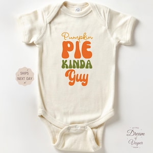 May include: Cream-colored baby bodysuit with the text "Pumpkin Pie Kinda Guy" in orange, green, and yellow. The bodysuit has short sleeves and a snap closure. A cute and cozy outfit for a baby.