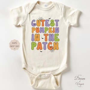 May include: Cream-colored baby bodysuit with the text "Cutest Pumpkin in the Patch" in orange, purple, green, and yellow. The design includes small spiderwebs and bats. A Halloween-themed infant outfit.