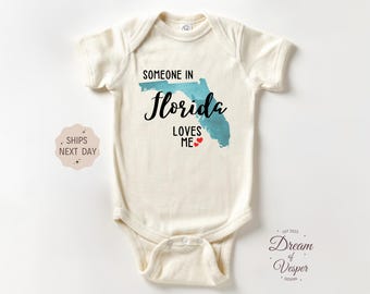 May include: Cream-colored baby onesie with the text "SOMEONE IN Florida LOVES ME" in black script and a teal Florida outline. Two small red hearts are below the text. The onesie is a short-sleeved style.