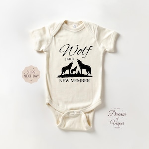 May include: Cream-colored baby onesie with a black wolf pack graphic. The design includes the words "Wolf pack" above three howling wolves and the text "NEW MEMBER" below. A cute baby outfit.