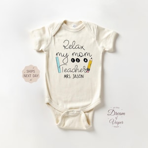 Peut inclure: Body de bébé crème avec l'inscription "Relax my mom is a Teacher" en écriture noire. Une règle bleue et un crayon jaune sont inclus. Le nom "MRS. JASON" est imprimé en dessous.