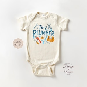 May include: Cream-colored baby bodysuit with the text "Tiny Plumber" in blue, featuring illustrations of plumbing tools. The design includes a wrench, faucet, and toolbox. The bodysuit is made of soft fabric.