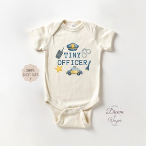May include: Cream-colored baby bodysuit with the words "TINY OFFICER" and illustrations of a police hat, handcuffs, a walkie-talkie, a star, a flashlight, and a police car. The bodysuit is short-sleeved.