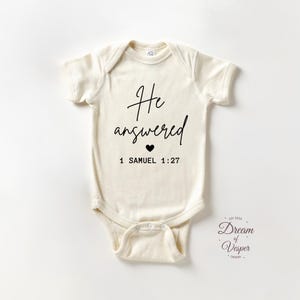 May include: Cream-colored baby onesie with short sleeves. The front features the text "He answered" in a handwritten font, a small black heart, and the scripture reference "1 Samuel 1:27". The Dream of Vesper Design logo is in the bottom right corner.