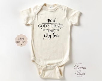 May include: Cream-colored baby onesie with the text "All of GOD'S GRACE in one tiny face" in black script. The onesie has short sleeves and a snap closure. A small sticker on the left says "SHIPS NEXT DAY".