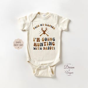 May include: Cream-colored baby bodysuit with the text "PACK MY DIAPERS" and "I'M GOING HUNTING WITH DADDY!" in brown and blue lettering. The design includes a deer and crossed rifles. A cute outfit for a baby.