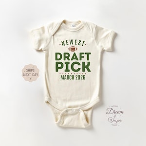 May include: Cream-colored baby onesie with the text "NEWEST DRAFT PICK MARCH 2026" in green and brown. The design includes a football graphic above the words "DRAFT PICK". The onesie has short sleeves and a snap closure.
