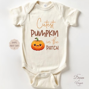 May include: Cream-colored baby bodysuit with the text "Cutest Pumpkin in the Patch" in brown and orange. A cartoon pumpkin graphic is also featured. The bodysuit is designed for infants.