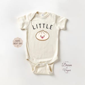 May include: Cream-colored baby bodysuit with the word "LITTLE" in black above a smiling dumpling illustration. The bodysuit is short-sleeved and has a snap closure at the bottom. The design is centered on the front.