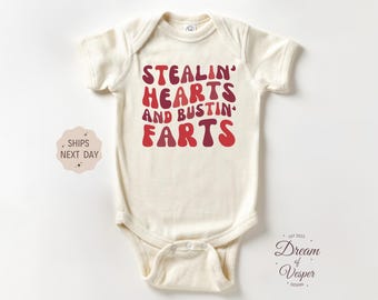 May include: Cream-colored baby onesie with the text "STEALIN' HEARTS AND BUSTIN' FARTS" in red and maroon lettering. The onesie has short sleeves and a snap closure. The design is playful and humorous.