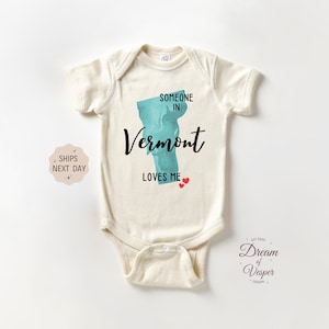 May include: Cream-colored baby bodysuit with the text "SOMEONE IN Vermont LOVES ME" in black script. A teal-colored outline of Vermont is in the center, with two small red hearts below the text. The bodysuit is short-sleeved.