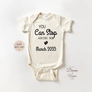May include: Cream-colored baby onesie with the text "YOU Can Stop ASKING NOW" in black. Below the text is a footprint graphic and the date "March 2025". The onesie is a short-sleeved garment.