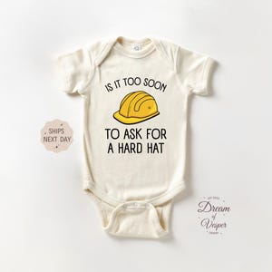 May include: Cream-colored baby onesie with the text "IS IT TOO SOON TO ASK FOR A HARD HAT" and a yellow construction helmet graphic. The onesie has short sleeves and a snap closure.