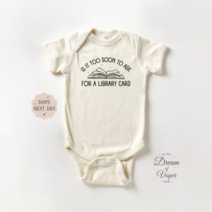 May include: A white baby onesie with black text that reads "Is it too soon to ask for a library card?" with a black and white illustration of an open book.