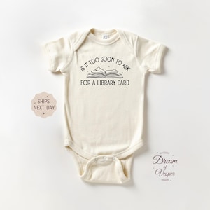 May include: Cream-colored baby bodysuit with short sleeves. The front features the text "IS IT TOO SOON TO ASK FOR A LIBRARY CARD" above an open book illustration. A small graphic with text is on the left side.