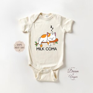 May include: Cream-colored baby bodysuit with a cartoon cat sleeping on a branch. The cat is white with orange spots and the text "MILK COMA" is below the cat. The bodysuit has short sleeves and a snap closure.