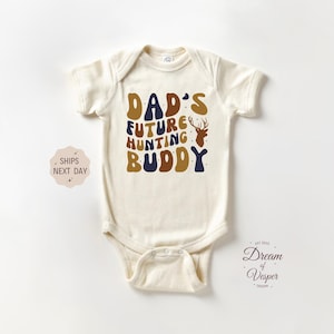 May include: Cream-colored baby onesie with the text "DAD'S FUTURE HUNTING BUDDY" in brown, navy, and gold lettering, along with a deer silhouette. A cute and stylish outfit for a baby.