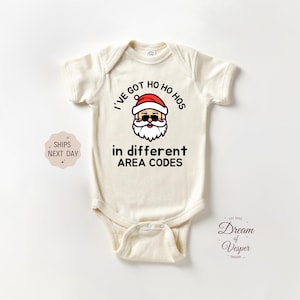 May include: Cream-colored baby bodysuit with the text "I'VE GOT HO HO HOS in different AREA CODES" and a Santa Claus graphic. The Santa graphic has sunglasses and a white beard. The bodysuit is short-sleeved.