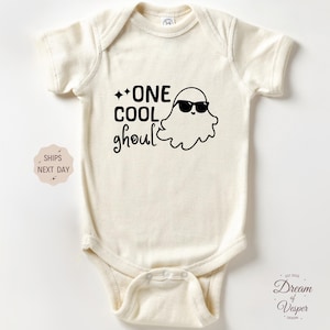 May include: Cream-colored baby bodysuit with a black graphic of a ghost wearing sunglasses and the words "ONE COOL ghoul". A fun Halloween-themed outfit for infants.