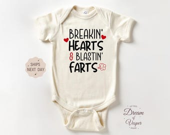 May include: Cream-colored baby onesie with the text "BREAKIN' HEARTS & BLASTIN' FARTS" in black, with red heart accents. The onesie has short sleeves and a snap closure. The design includes a small flower graphic.