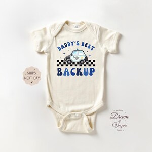 May include: Cream-colored baby bodysuit with the text "DADDY'S BEST BACKUP" in blue. A police car graphic is above the word "BACKUP". The bodysuit has short sleeves and a snap closure.