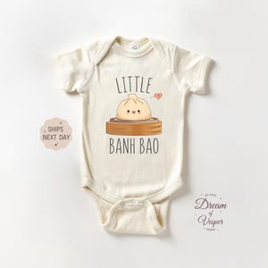 May include: Cream-colored baby onesie with the words "Little Banh Bao" and a cartoon dumpling illustration. The design includes a small heart and is printed on a soft, short-sleeved garment.