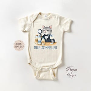 May include: Cream-colored baby bodysuit with a whimsical illustration of a cat sommelier. The cat, wearing a tuxedo, holds a magnifying glass and a glass of milk. The text "MILK SOMMELIER" is below the illustration.