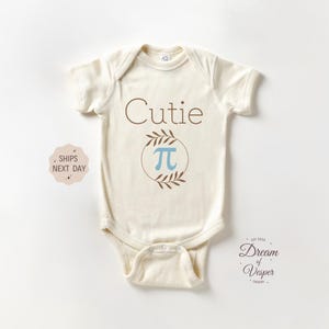 May include: A cream-colored baby onesie with the text "Cutie" and a circle with the Greek letter "pi" inside. The circle is surrounded by brown leaves.