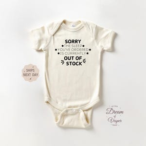May include: Cream-colored baby bodysuit with the text "SORRY THE SLEEP YOU'VE ORDERED IS CURRENTLY OUT OF STOCK" in black. The bodysuit has short sleeves and a snap closure. A small sticker says "SHIPS NEXT DAY".