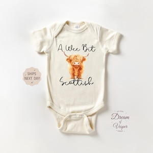 May include: Cream-colored baby bodysuit with the text "A Wee Bit Scottish" and a watercolor illustration of a Highland cow. The bodysuit has short sleeves and a snap closure.
